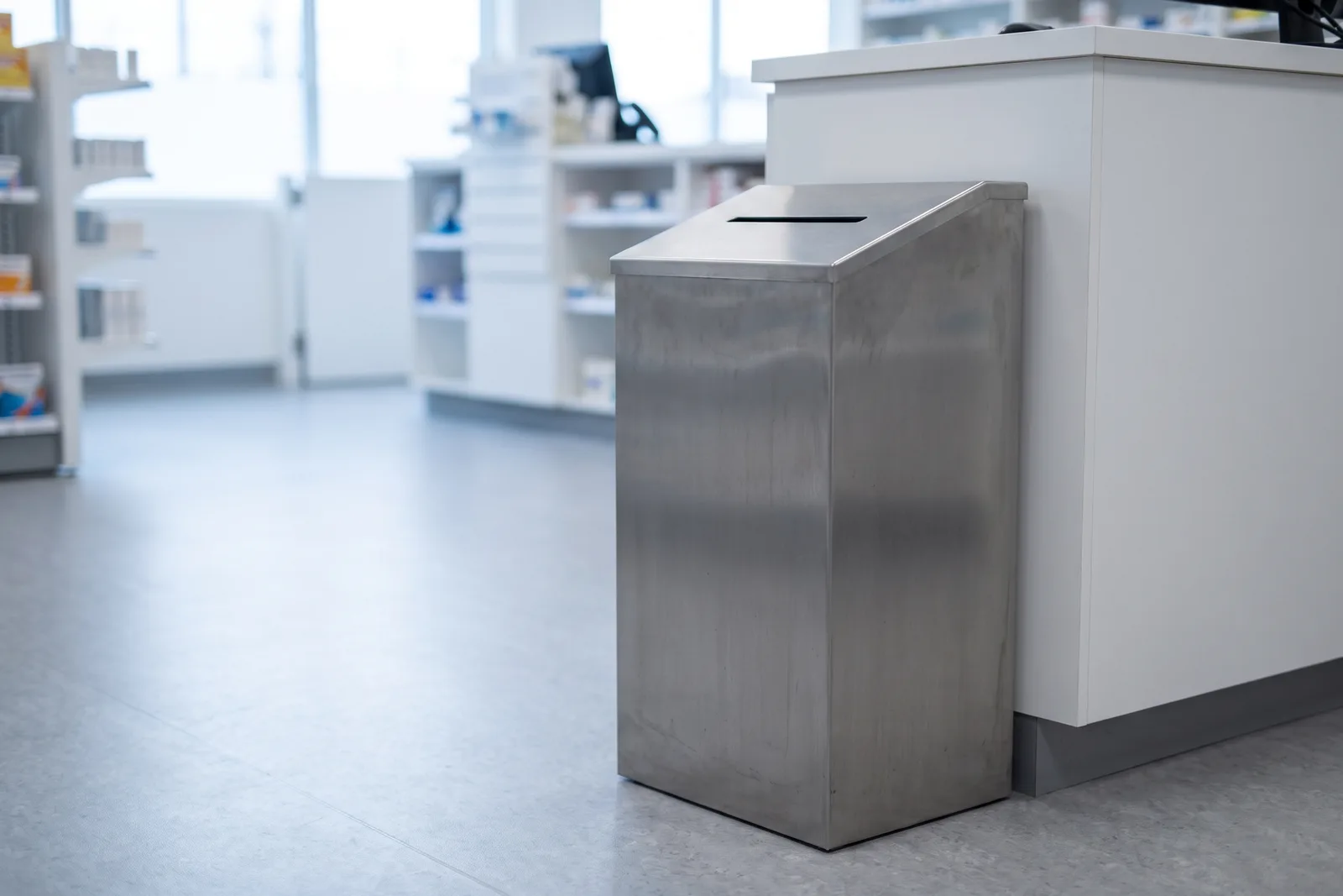A steel medication drop-off bin standing beside a pharmacy counter